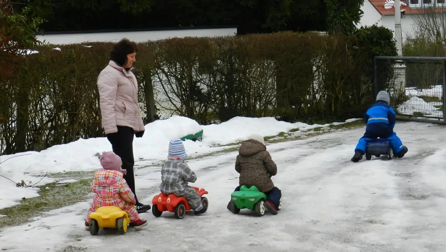 Tagesmütter Augsburg-Land e.V. - Kita-Trägerverband - Ein Erwachsener beobachtet drei kleine Kinder, die mit Spielzeugautos auf einem verschneiten Weg fahren.