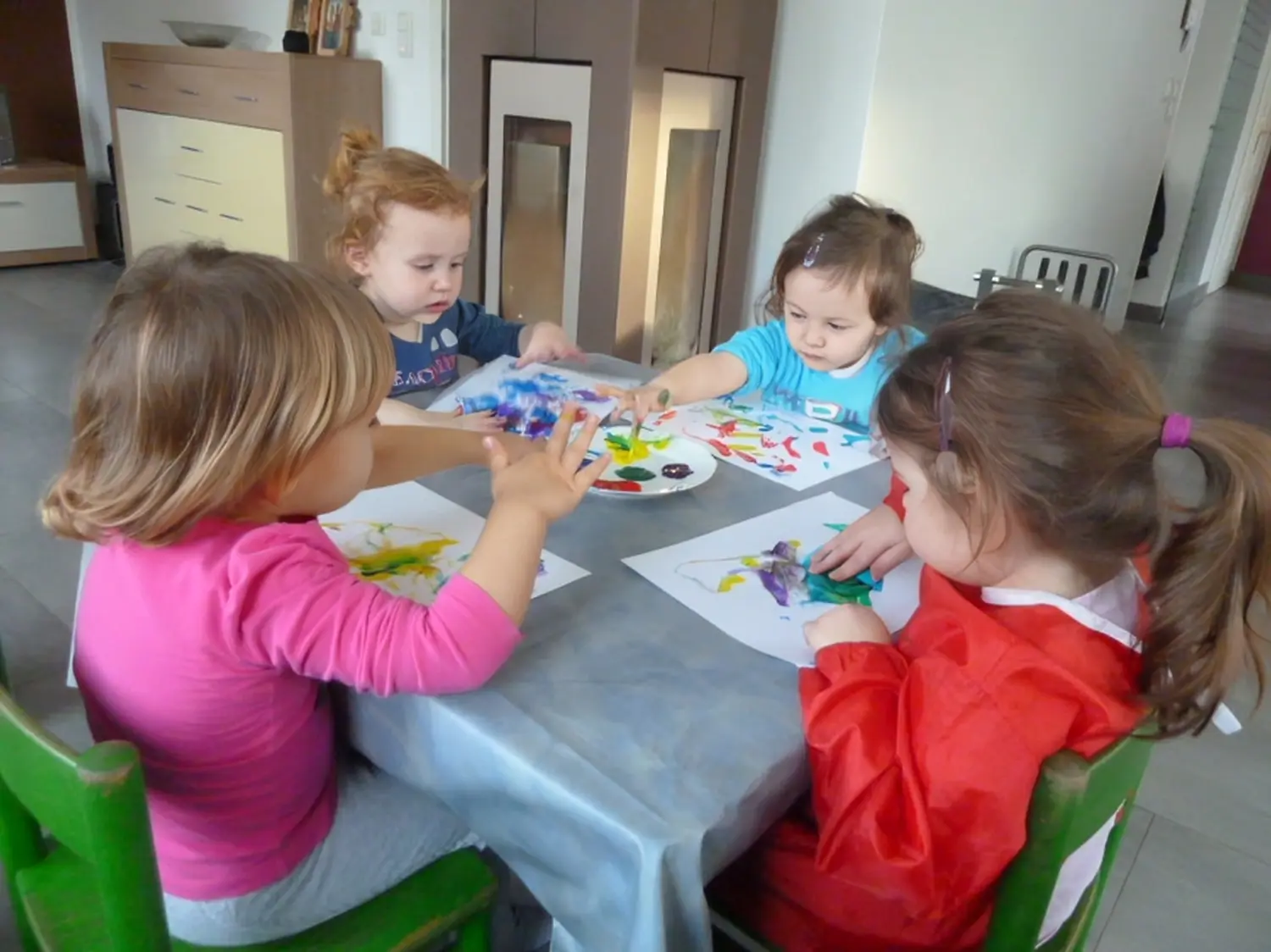 Tagesmütter Augsburg-Land e.V. - Kita-Trägerverband - Vier kleine Kinder sitzen um einen Tisch und malen mit den Fingern auf Papier.