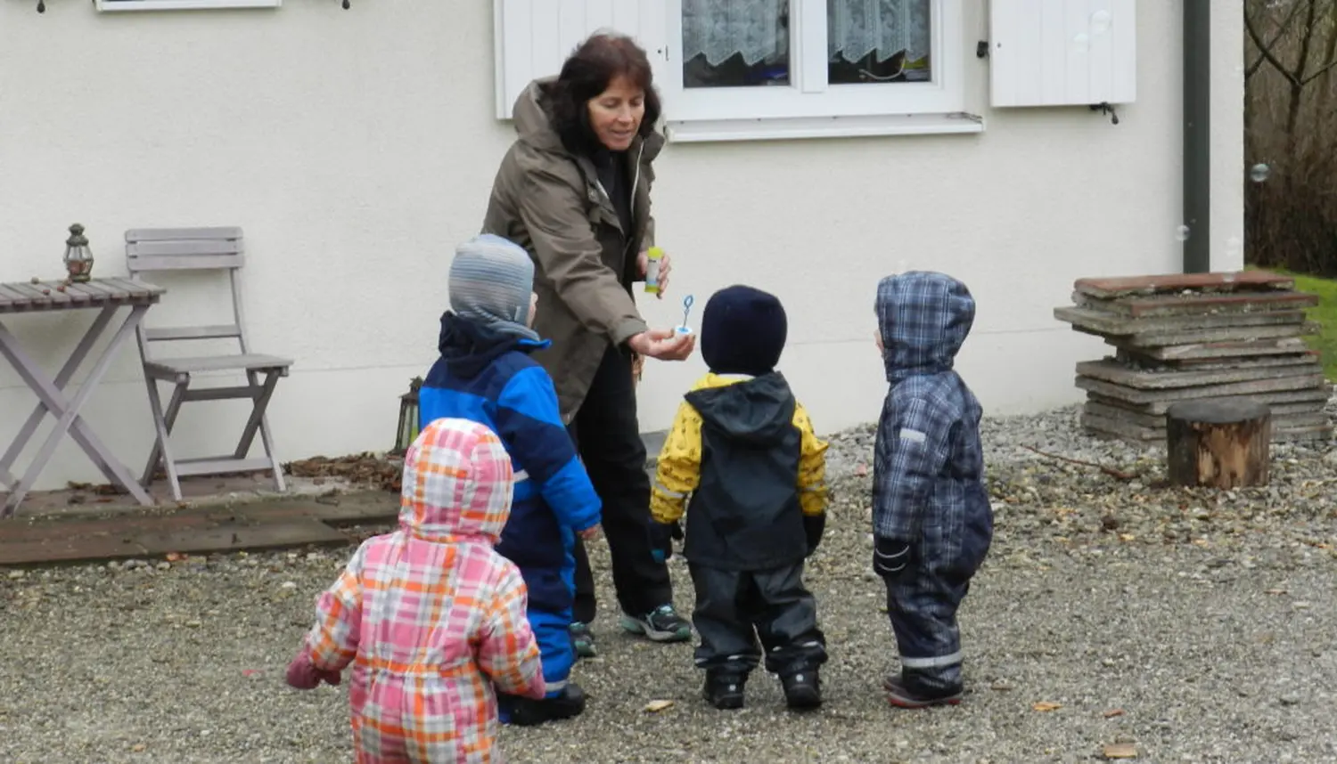 Tagesmütter Augsburg-Land e.V. - Kita-Trägerverband - Eine erwachsene Frau überreicht vier kleinen Kindern in Wintermänteln im Freien ein Spielzeug.