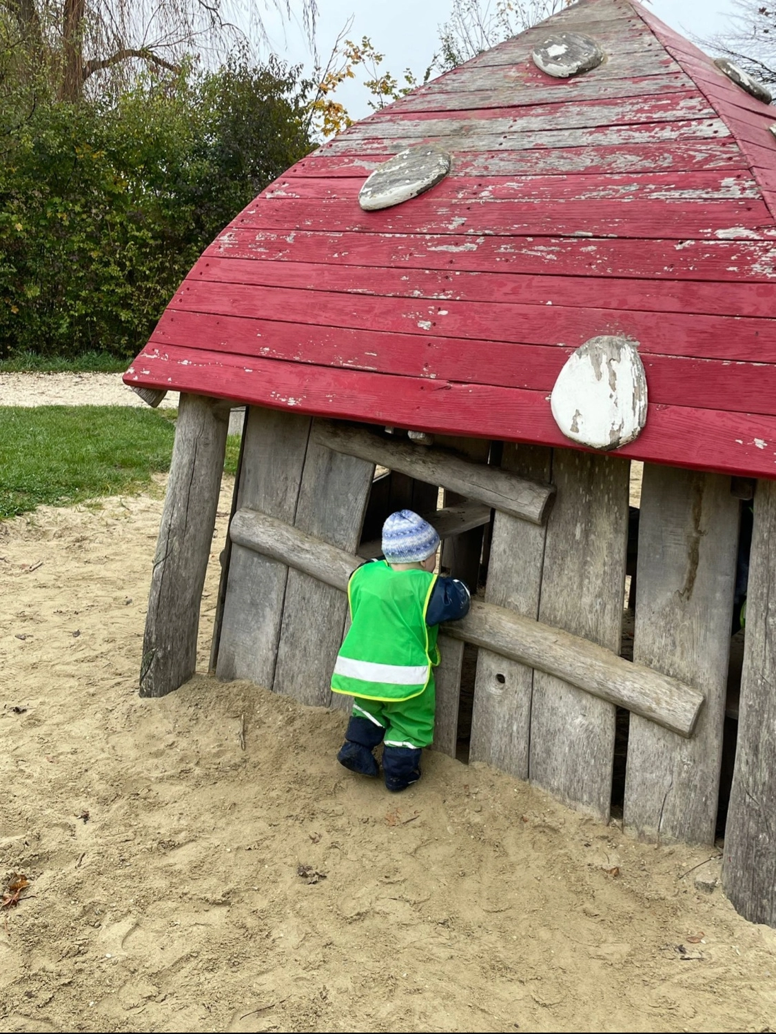 Tagesmütter Augsburg-Land e.V. - Kita-Trägerverband - Ein Kleinkind in einer leuchtend grünen Sicherheitsweste schaut durch ein Fenster eines Holzspielhauses auf einem Sandspielplatz.